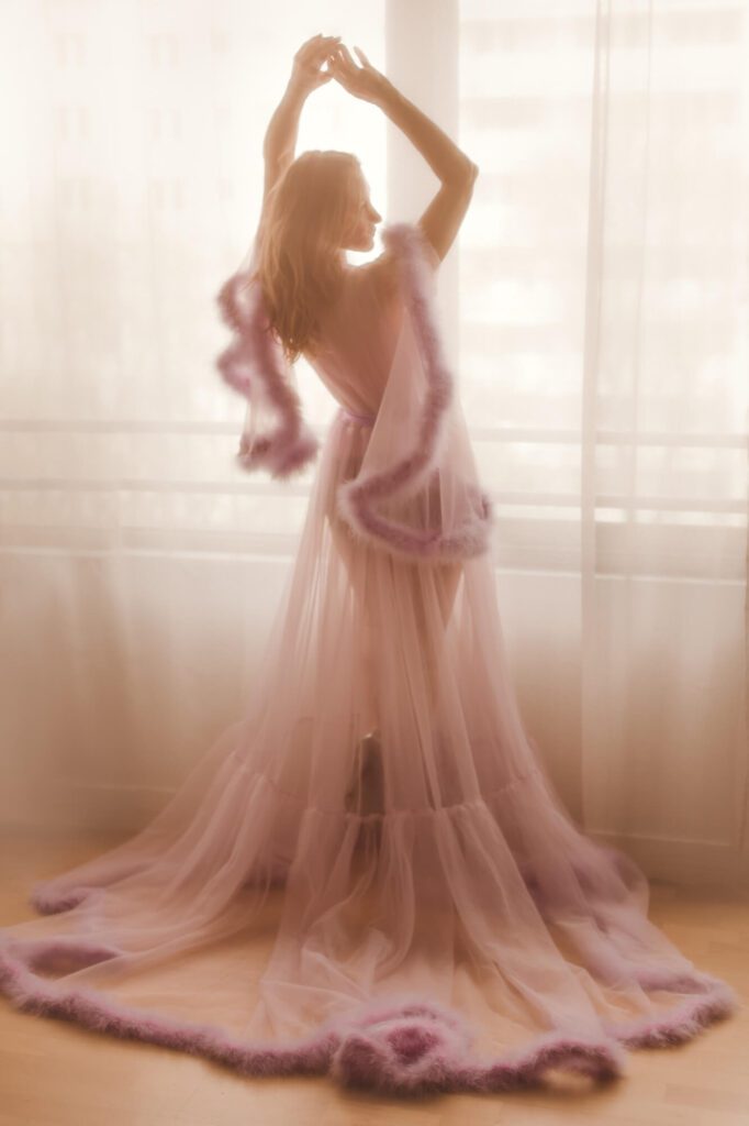 Kobieta z podniesionymi do góry rękoma stoi przy oknie w liliowym peniuarze obszytym piórami.