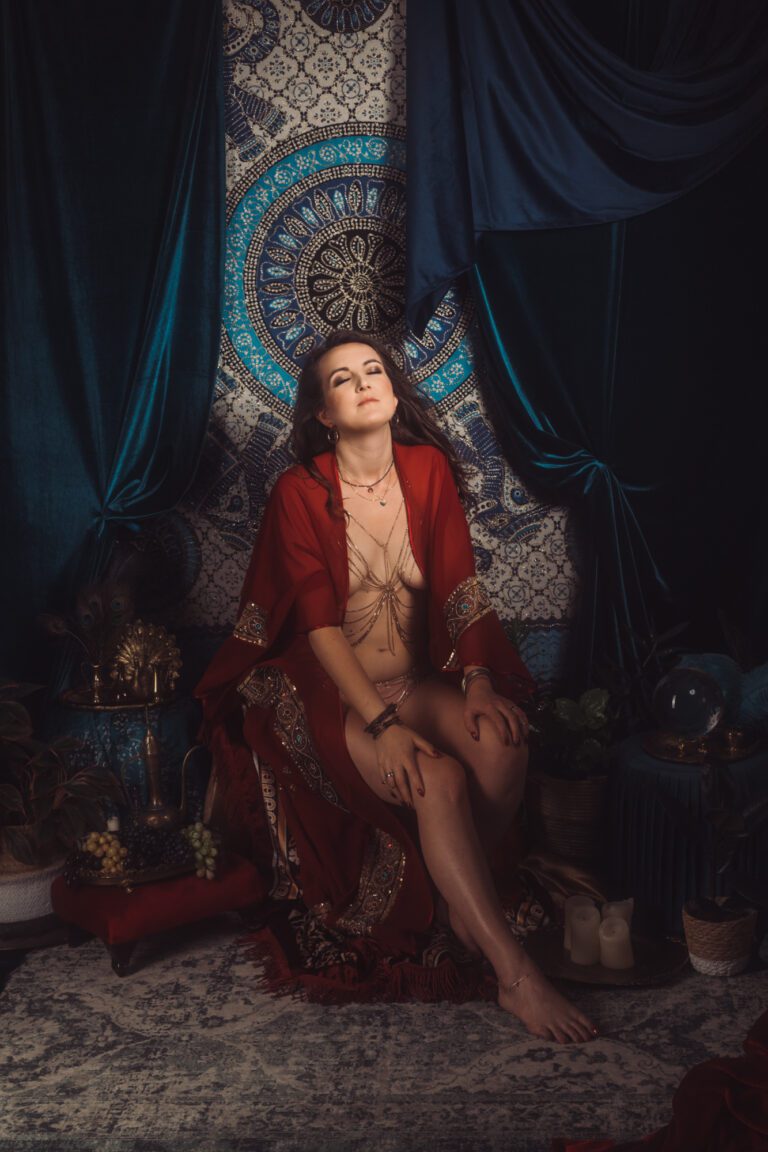 Kobieta ubrana w bordowy peniuar siedzi na tle ozdobnego gobelinu z niebiesko-złotymi wzorami. Otacza ją aksamitna draperia, rośliny, świece i owoce. Ma zamknięte oczy i dłonie na kolanach.