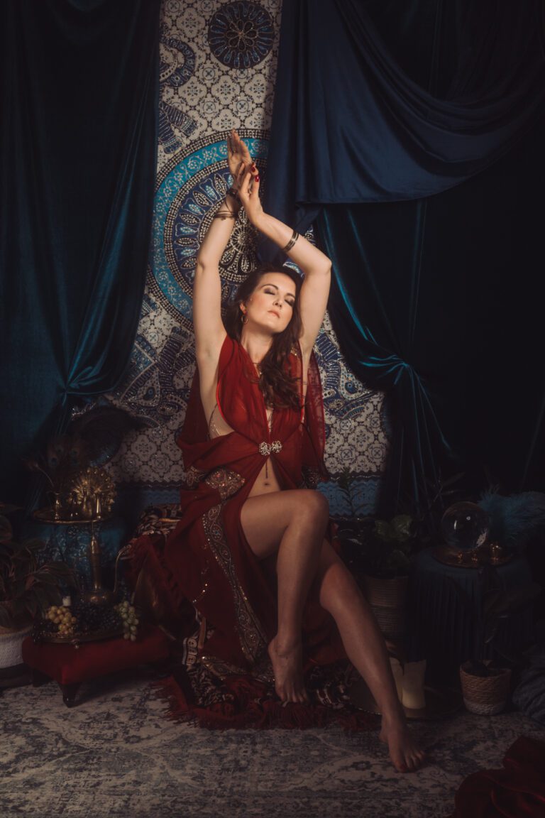 Kobieta ubrana w bordowy peniuar siedzi na tle ozdobnego gobelinu z niebiesko-złotymi wzorami. Otacza ją aksamitna draperia, rośliny, świece i owoce. Ma zamknięte oczy i ręce ma skierowane w górę.