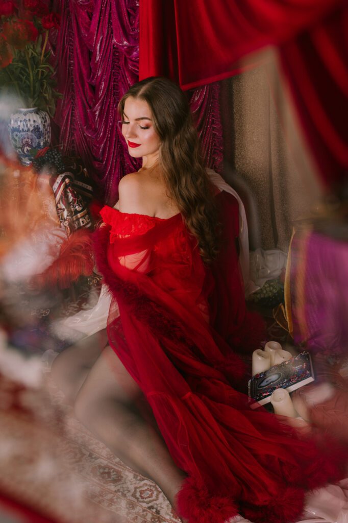 Kobieta z długimi brązowymi włosami siedzi na podłodze i ogląda się przez ramię w marzycielskim, słabo oświetlonym otoczeniu z luksusowymi czerwonymi zasłonami. Ma na sobie przezroczystą czerwoną szatę odsłaniającą ramiona, jest otoczona ozdobnymi dekoracjami, świecami.