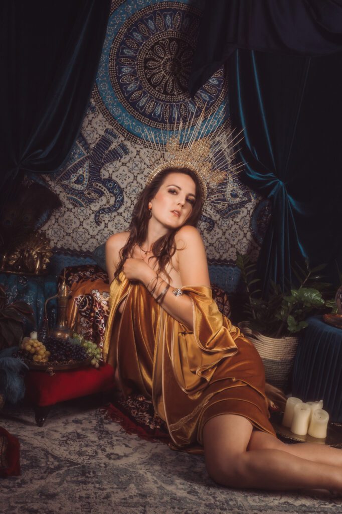 Kobieta ubrana w złotą sukienkę i koronę wykonaną z cienkich, kolczastych promieni siedzi na tle ozdobnego gobelinu z niebiesko-złotymi wzorami. Otacza ją aksamitna draperia, rośliny, świece i owoce. Z pewnością patrzy w kamerę.