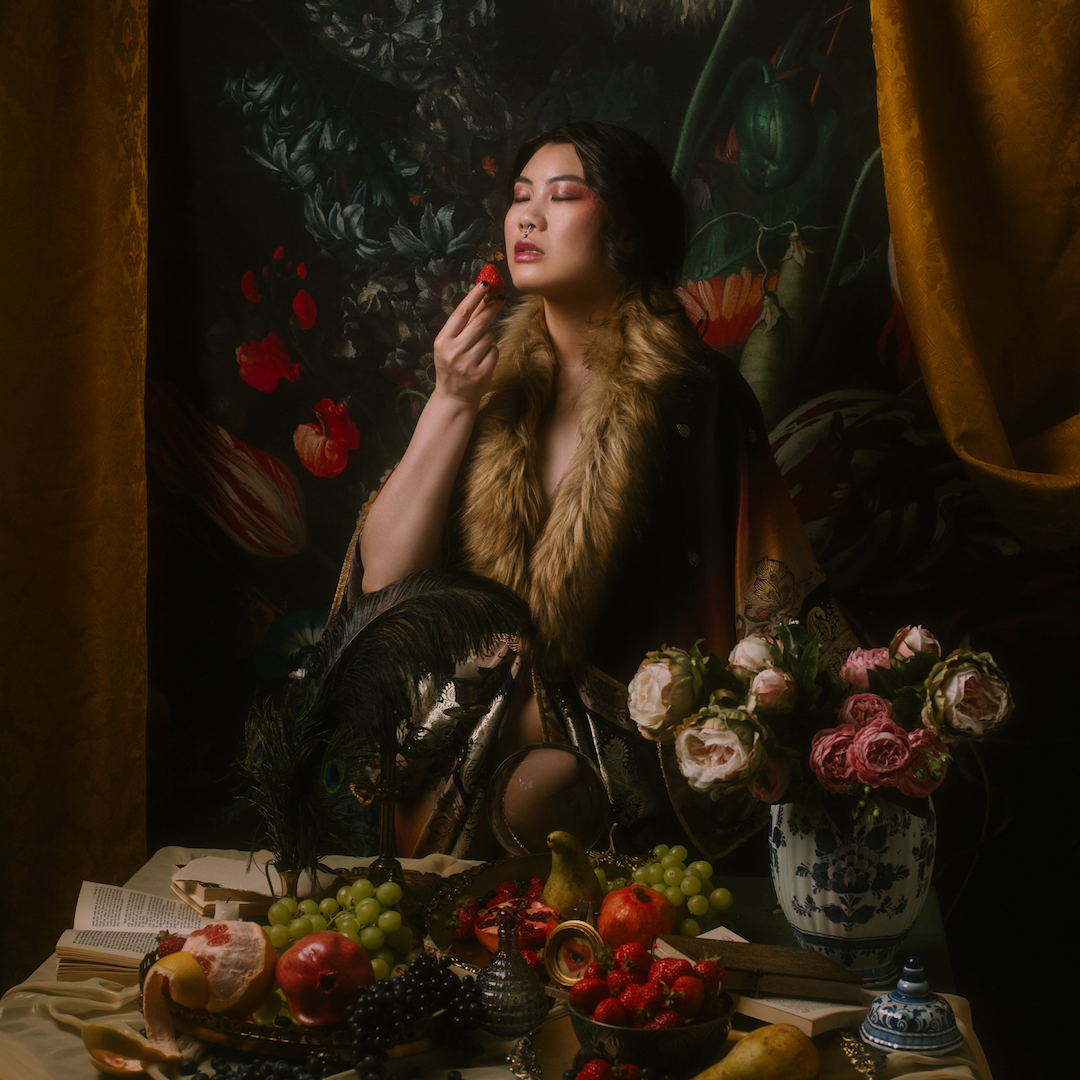 Kobieta ubrana w luksusową szatę obszytą sztucznym futrem z gracją trzyma w ustach truskawkę, otoczona wystawną ucztą owoców i kwiatów na bogato zdobionym stole. Tło jest ciemne, z odważnymi kwiatowymi wzorami, przywołującymi klasyczną, wystawną atmosferę.