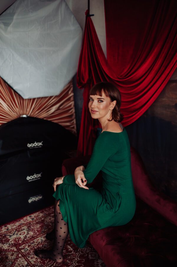 Kobieta w zielonej sukience uśmiecha się i patrzy w obiektyw, siedzi na bordowej sofie.