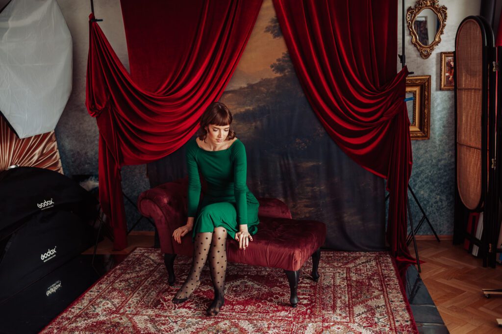 Kobieta w zielonej sukience siedzi na bordowym szezlongu w pokoju ozdobionym czerwonymi aksamitnymi zasłonami i wystrojem w stylu vintage. Patrzy w dół. W tle namalowany pejzaż oraz wzorzysty dywanik na podłodze.
