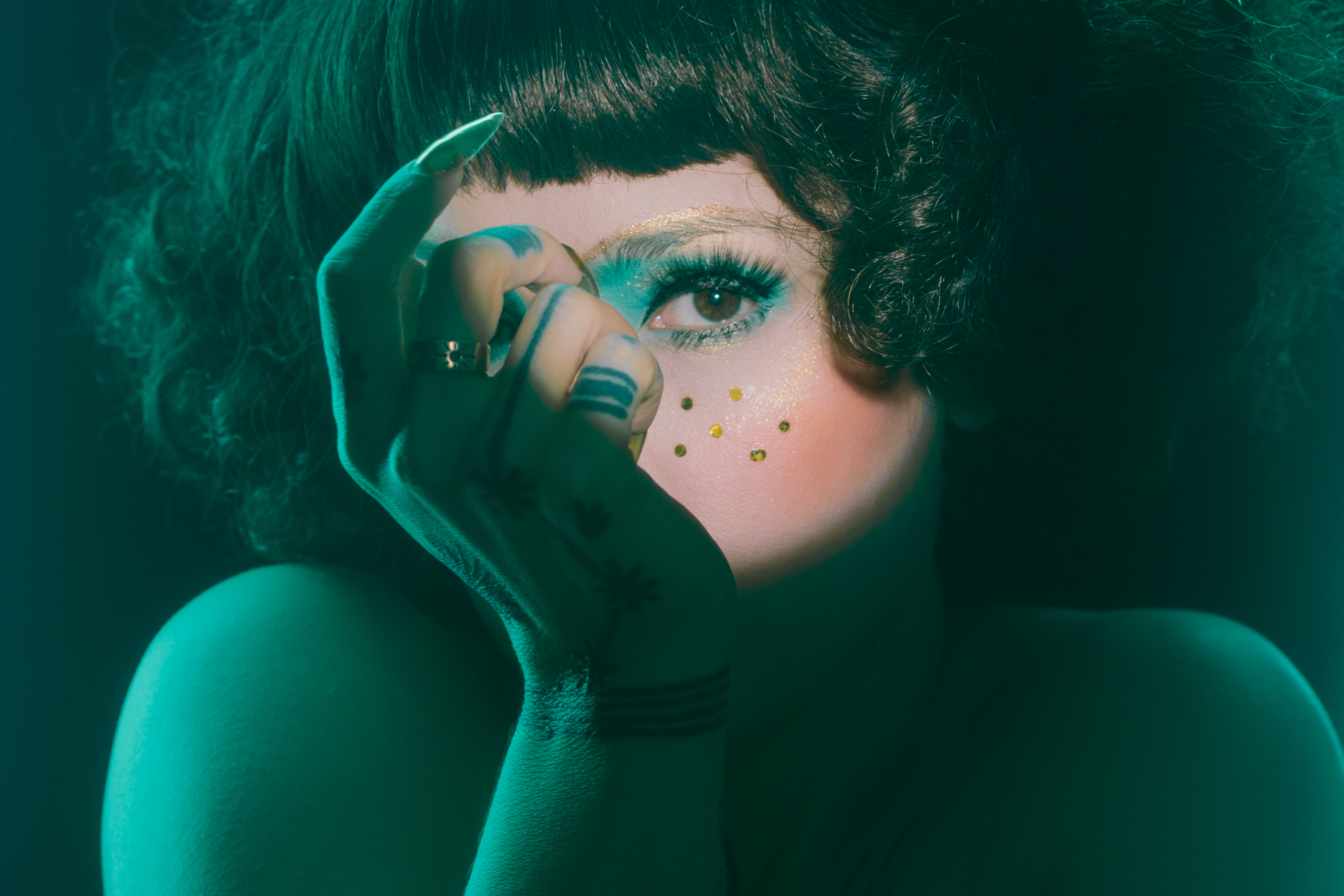 Osoba z fryzurą na kręcony bob i efektownym makijażem, zawierającym niebieski cień do powiek i złote kropki na twarzy, częściowo zasłania twarz jedną ręką. Oświetlenie rzuca na scenę zielonkawy odcień, tworząc dramatyczną i tajemniczą atmosferę.