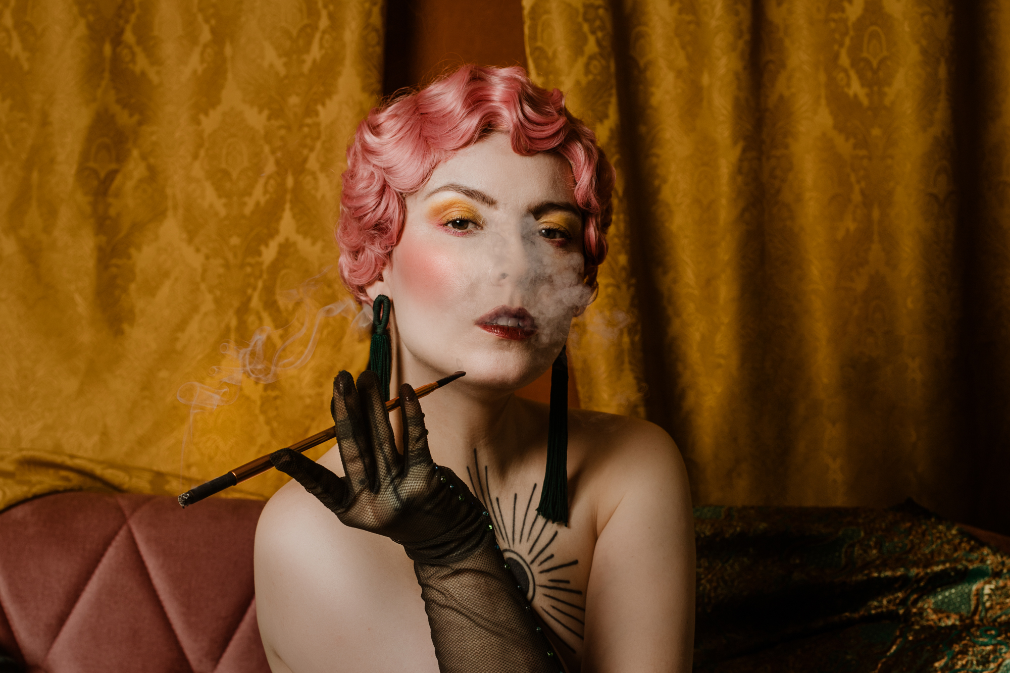 Kobieta z różowymi włosami w stylu lat 20. pali papierosa z fifką. Ma czarne rękawiczki.