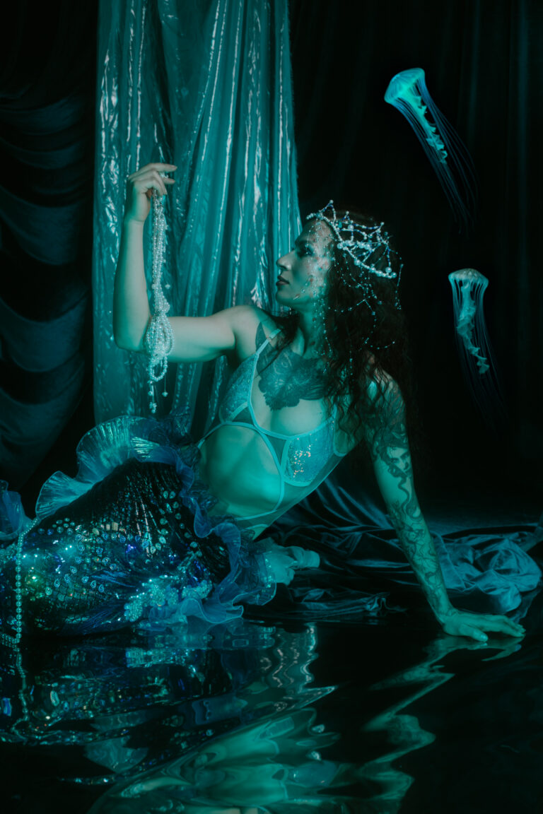 Kobieta z ogonem syreny i diademie z pereł w turkusowym świetle.